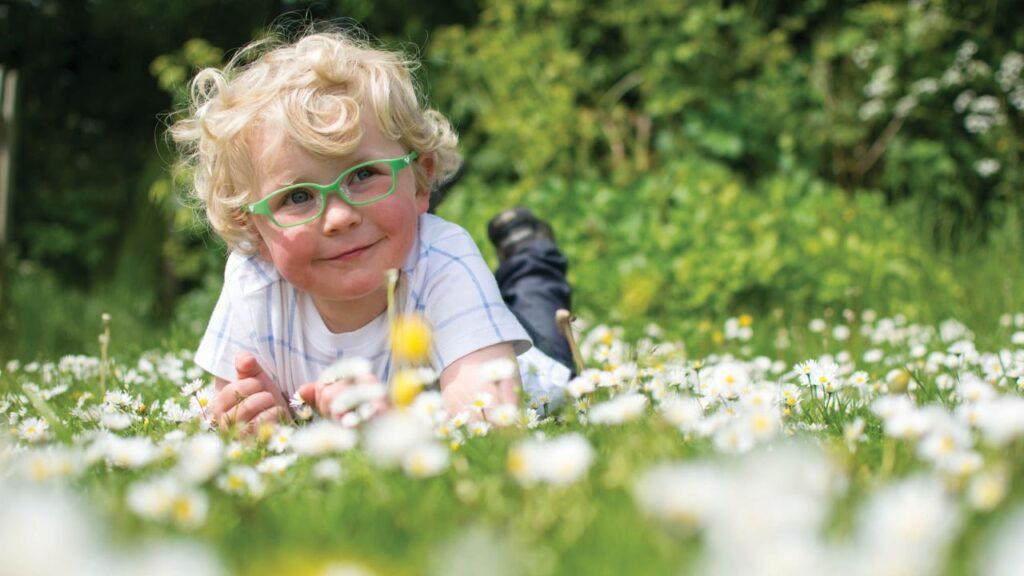 Toddler lying in a flowery meadow with green glasses on. EK Eyewear Belfast.